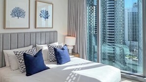 Quarto de hotel moderno com uma cama grande, duas obras de arte emolduradas na parede, uma mesa de cabeceira com abajur e vista para edifícios altos através de grandes janelas.