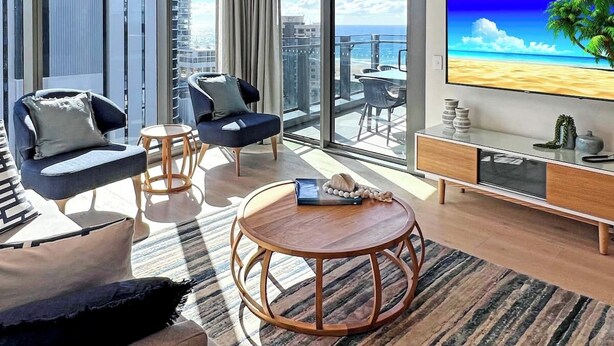 Uma sala de estar moderna com uma televisão grande, assentos confortáveis, uma mesa de centro redonda e vista para o oceano.