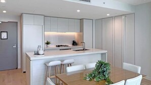 Cozinha moderna com armários brancos, uma ilha central e uma área de refeições com mesa de madeira e cadeiras brancas.