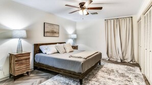 Un dormitorio con una cama de madera, dos lámparas de noche, un ventilador de techo, un ventanal con cortinas y una alfombra en el suelo.