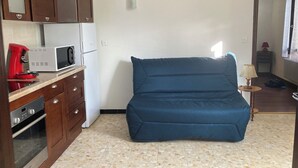 Ett kompakt kök med mikrovågsugn, kaffebryggare och ugn. I vardagsrummet finns en blå sittpuff.