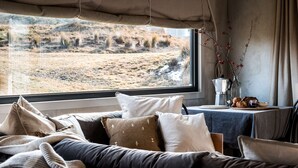 Une pièce confortable avec une grande fenêtre, un lit garni de plusieurs oreillers, une petite table avec une cafetière et des pâtisseries, et une vue sur un paysage aride à l’extérieur.