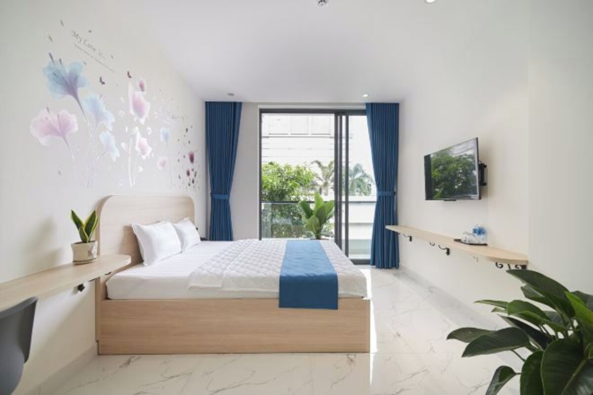 A modern hotel room with a large bed, a TV, a balcony with curtains, and a wall with floral artwork.