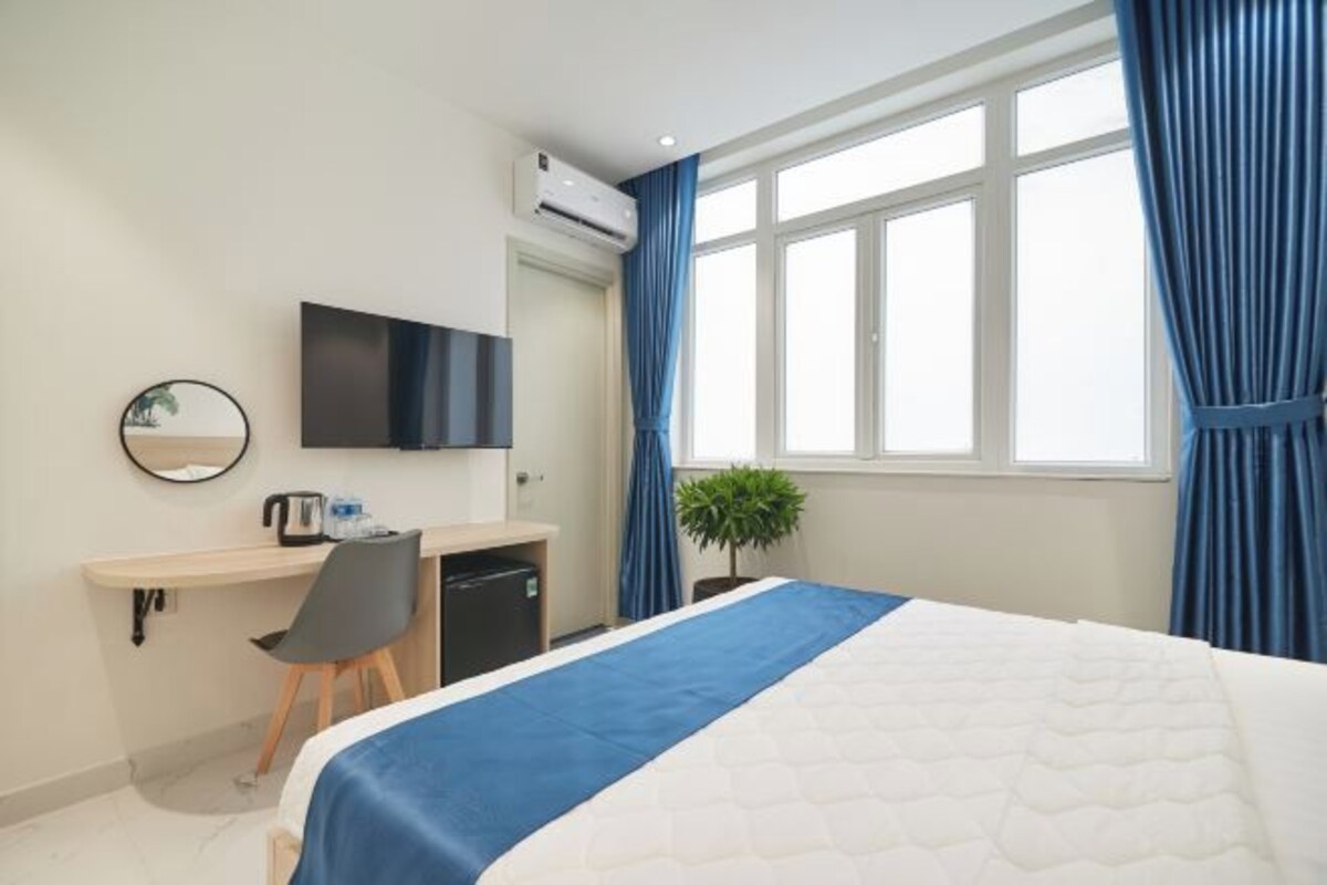 A hotel room with a bed, a desk with a chair, a television, a small plant, and a window with curtains.
