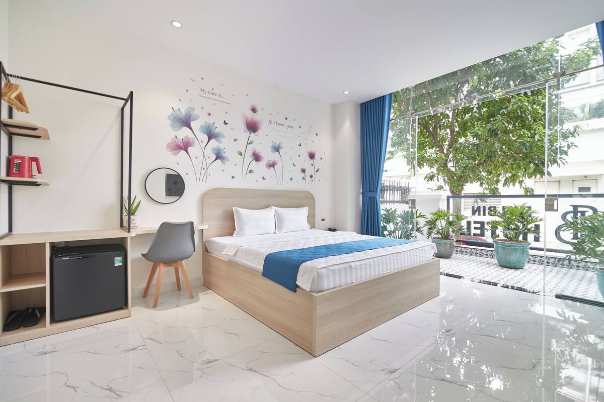 Ein modernes Hotelzimmer mit Bett, Schreibtisch, kleinem Kühlschrank und einem großen Fenster mit Blick ins Freie.