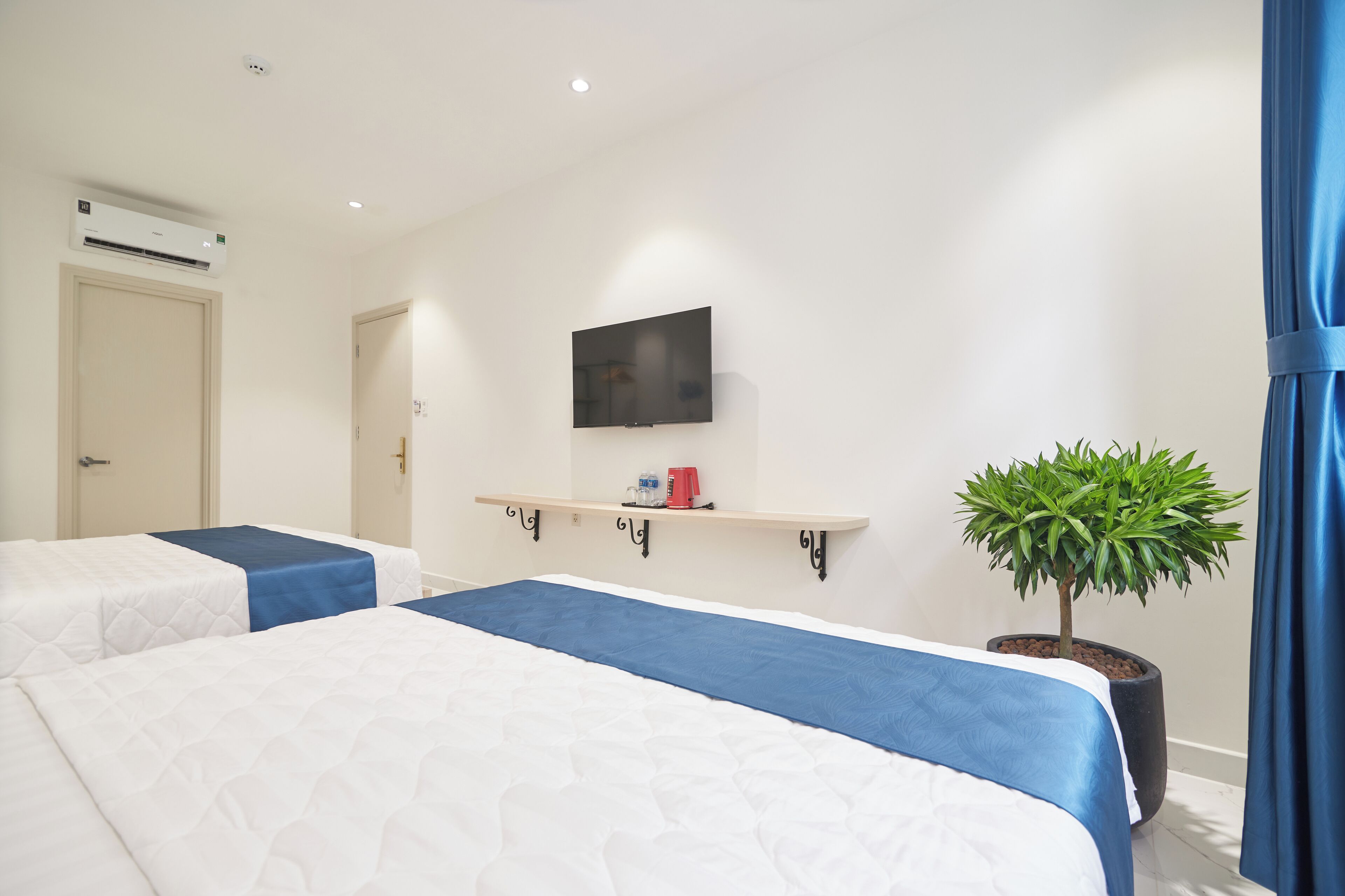Une chambre d’hôtel avec deux lits, un téléviseur fixé au mur, une étagère avec une plante et un appareil de climatisation.