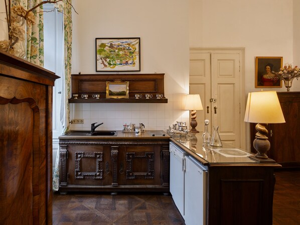 A traditional kitchen with wooden cabinetry, a built-in sink, and a countertop. There is a framed painting above the sink, a door with a mirror, and a vase with flowers on the right side.