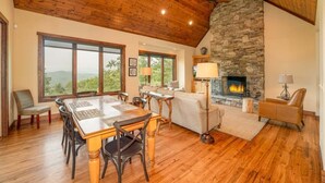 Una habitació àmplia amb sostre de fusta, llar de pedra, finestres grans i zona de dinar.