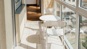 Un balcón con una mesa y sillas blancas, vistas a edificios, y una puerta que da a un espacio interior.
