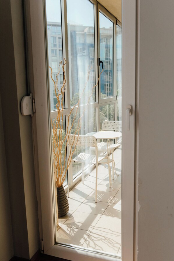 A view through a glass door into a balcony with a white table and chairs, a potted plant, and a cityscape in the background.