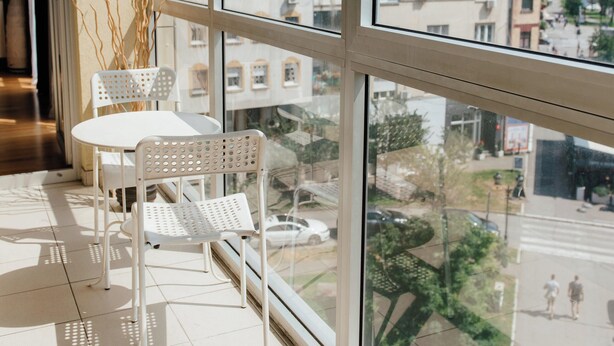 Un balcón con una mesa y sillas blancas, ventanas amplias y vistas a edificios y árboles.