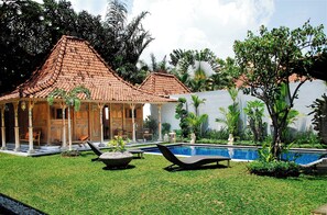 Relax in a serene outdoor setting with a traditional-style building, lounge chairs, and a swimming pool surrounded by lush greenery.