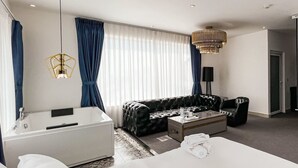 Quarto de hotel moderno com sofá, mesa de centro, escrivaninha, TV de tela plana, luminária e uma grande janela com cortinas.