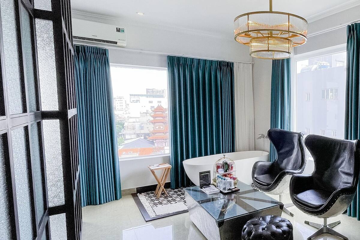 A modern room with a glass table, two black leather chairs, a black ottoman, a small side table, a black speaker, and teal curtains.