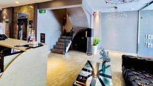 Um saguão de hotel com balcão de recepção, escada, lustre moderno e área de estar.