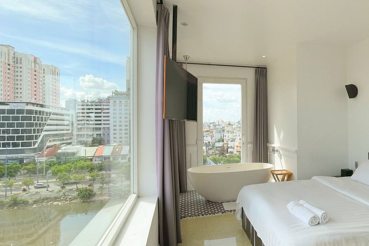 A hotel room with a large bed, a small side table, a nightstand, a window with a view of the city, and a bathroom with a bathtub.