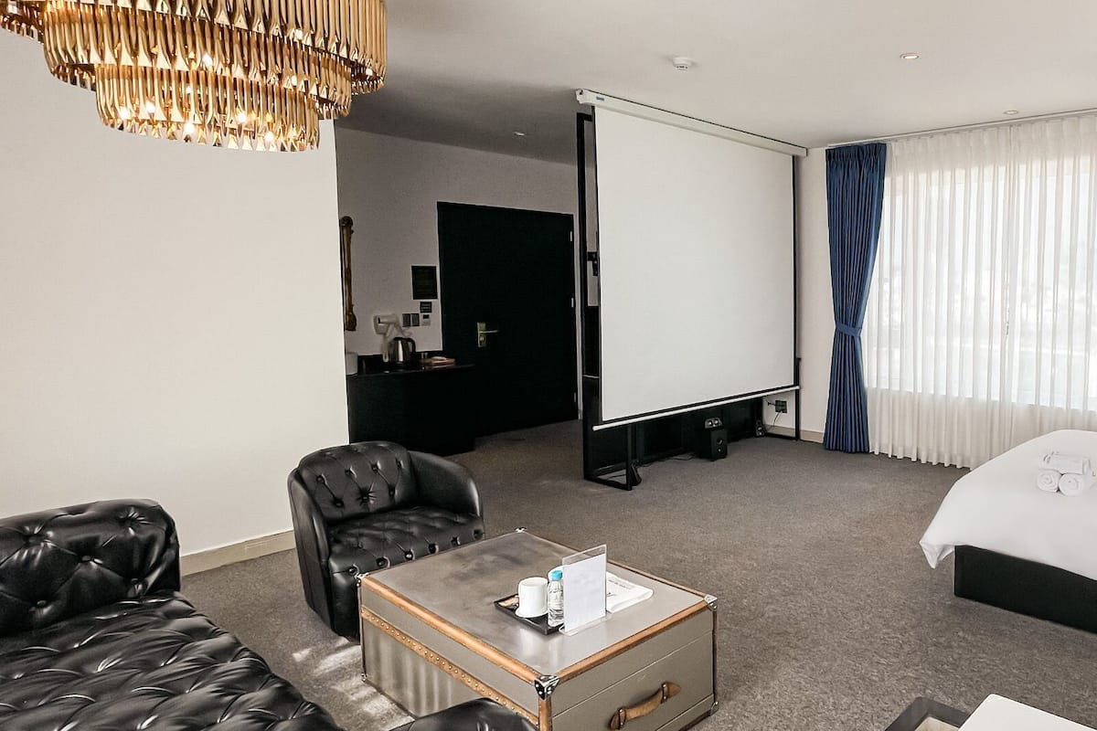 A hotel room with a bed, a leather sofa, a coffee table, a desk with a computer, and a projector screen.