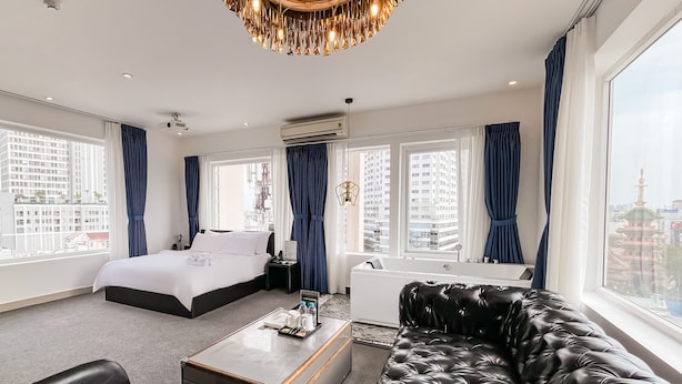 Quarto de hotel moderno com uma cama grande, um sofá de couro, uma mesinha e vista da cidade através de grandes janelas.