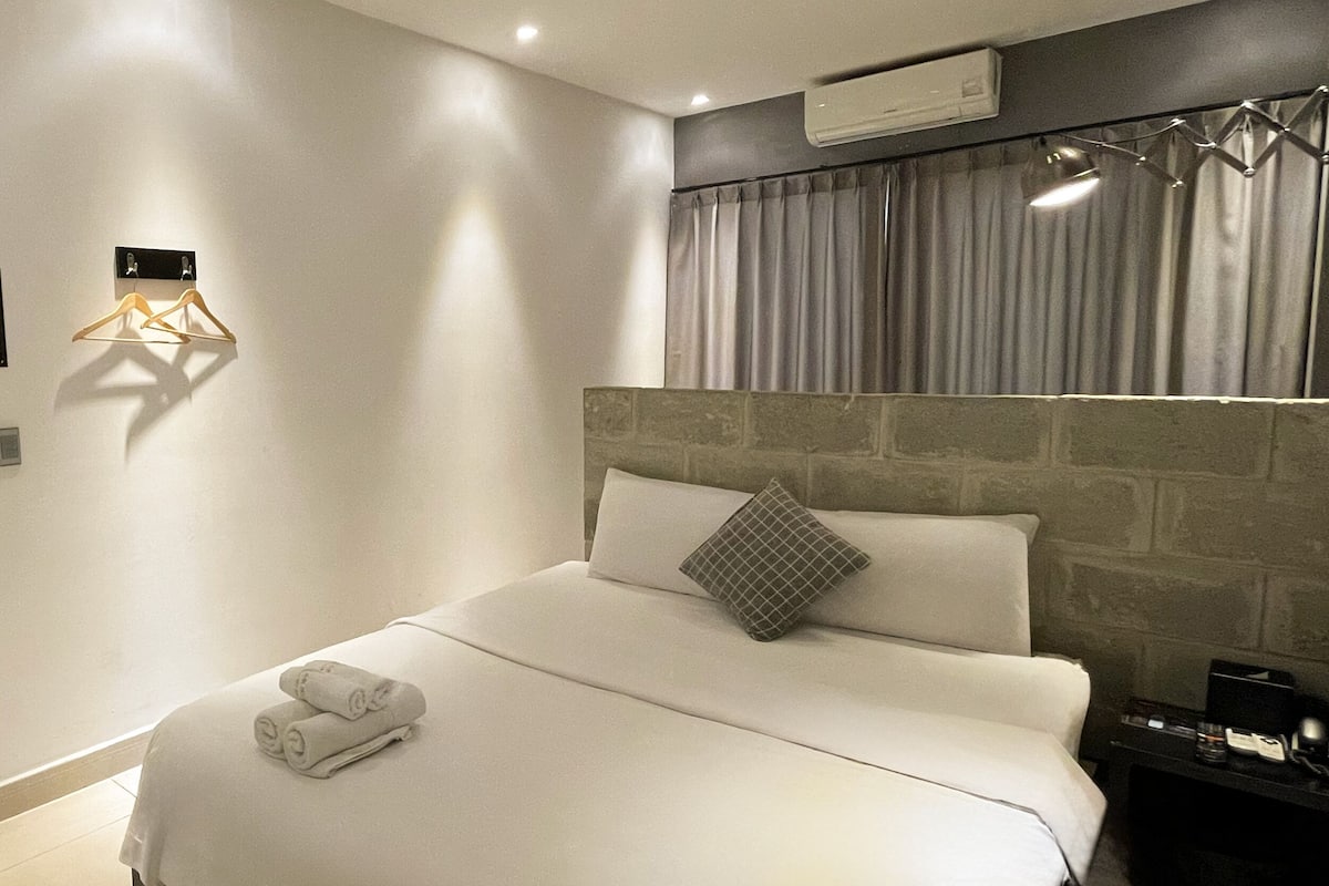 A modern hotel room with a bed, a black door, a wall-mounted light fixture, and a window with curtains.