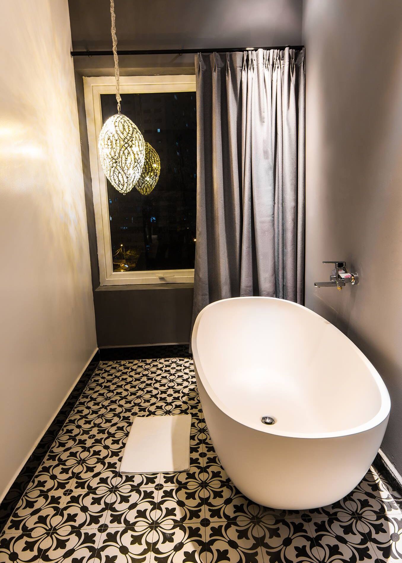 Un baño moderno con una bañera independiente, baldosas de piso estampadas, una lámpara colgante y una ventana con cortinas.