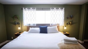 Una cama bien hecha con sábanas blancas, un cojín decorativo azul y un suave plaid blanco. Hay dos lámparas de noche a cada lado de la cama, y una ventana con cortina estampada.