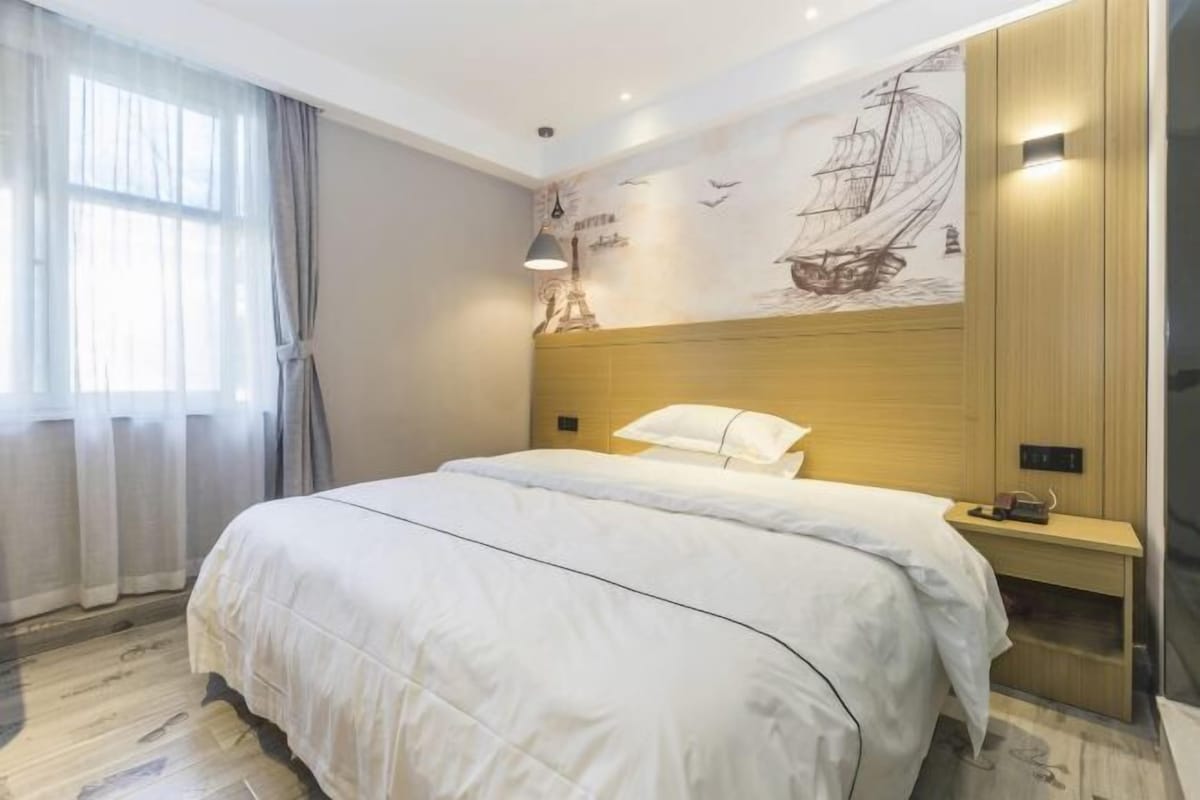 A modern hotel room with a large bed, a wooden headboard, a nightstand, a wall mural, and a window with curtains.