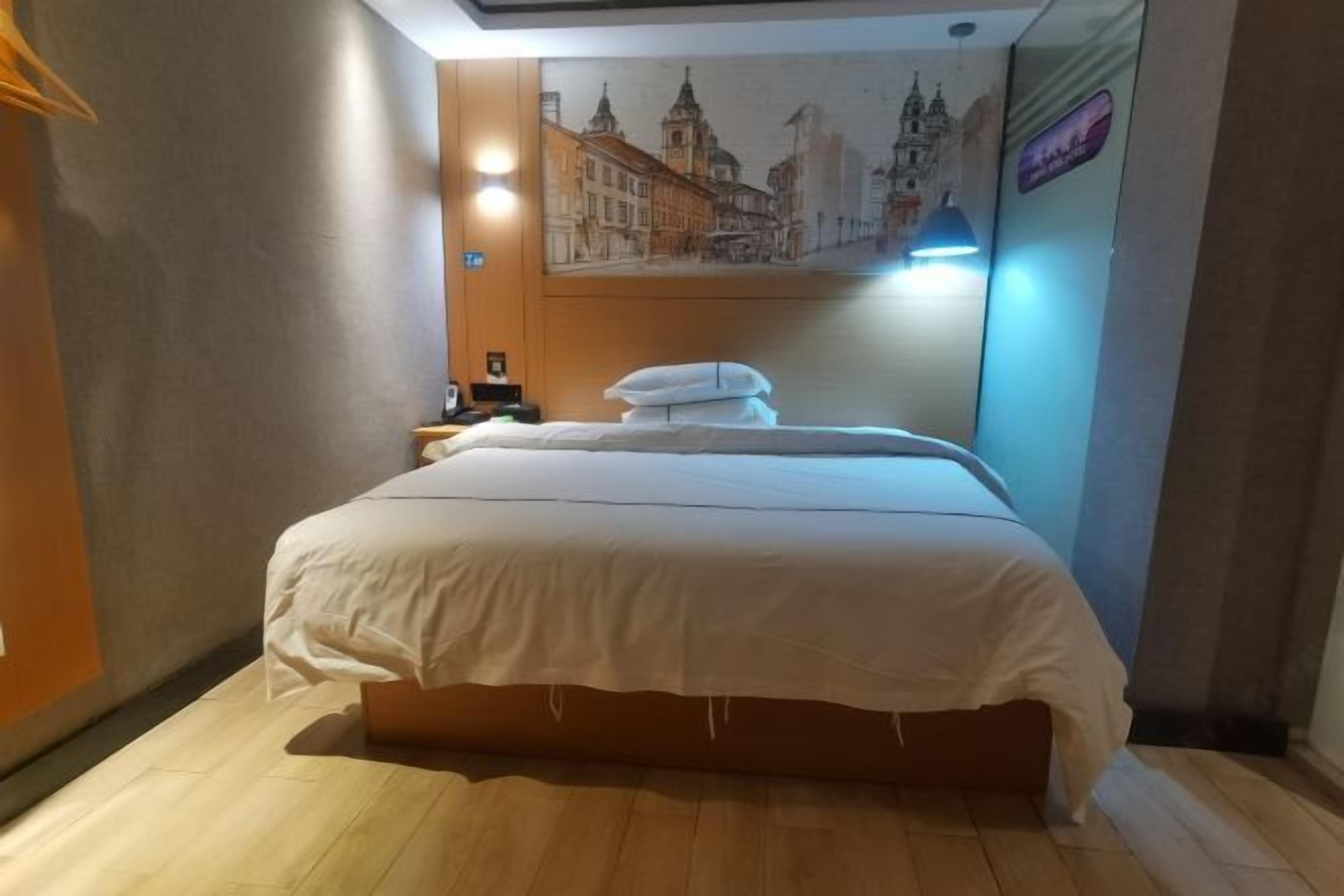 A hotel room with a large bed, a nightstand, a wall-mounted lamp, and a painting of a cityscape.