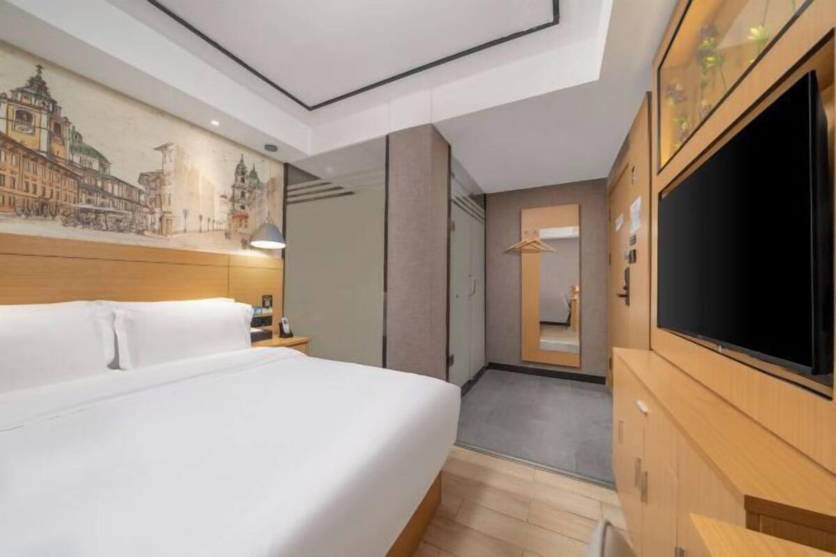 A hotel room with a bed, a television, a mirror, and a wall with a cityscape mural.