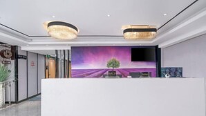 Un espace d’accueil avec un comptoir de réception blanc, une grande fresque murale aux couleurs violettes et roses représentant un arbre et un champ, et deux lustres.