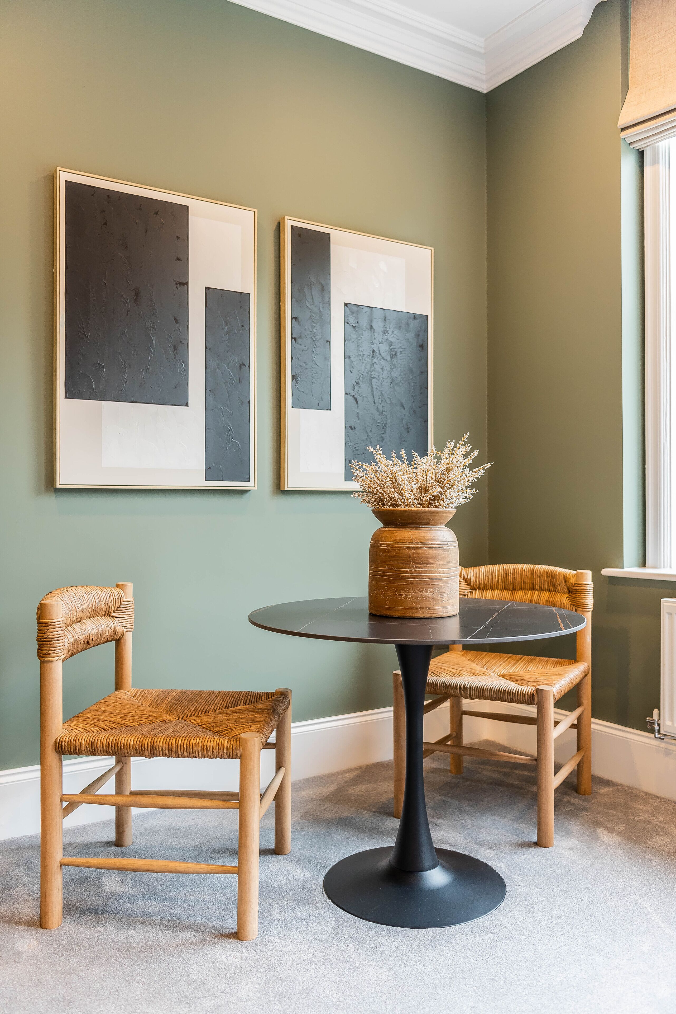 A room with two framed abstract paintings, a round table with a vase of dried flowers, and two wicker chairs.