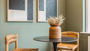 A room with two framed abstract paintings, a round table with a vase of dried flowers, and two wicker chairs.