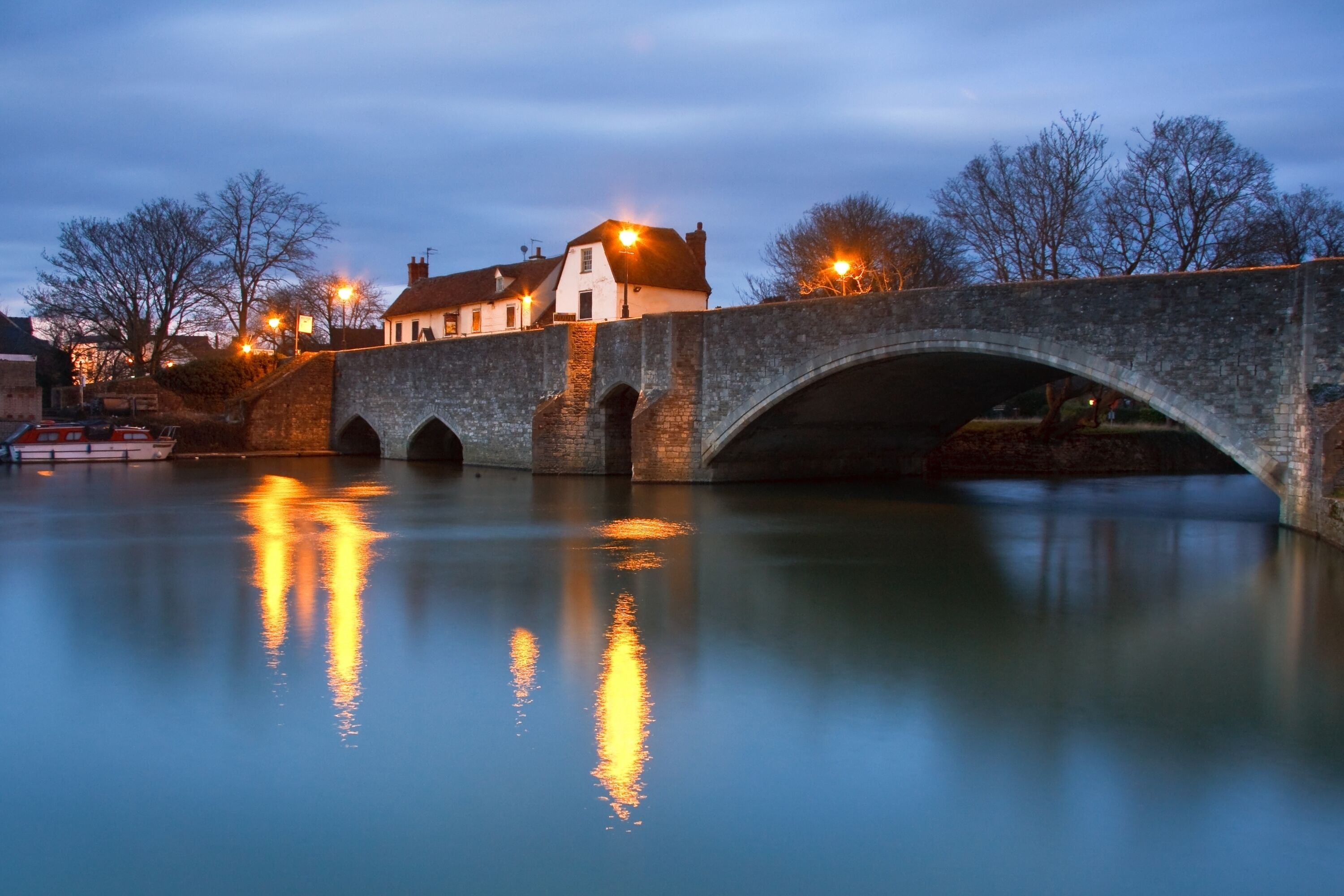 A stone bridge over a calm river at dusk with a house on the left side.