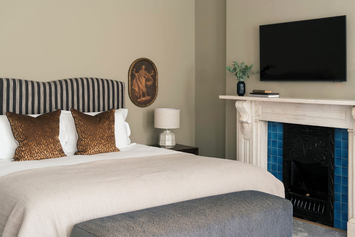 Een verzorgde slaapkamer met een groot bed, een televisie boven een open haard, en een klein tafeltje met een lamp en boeken.