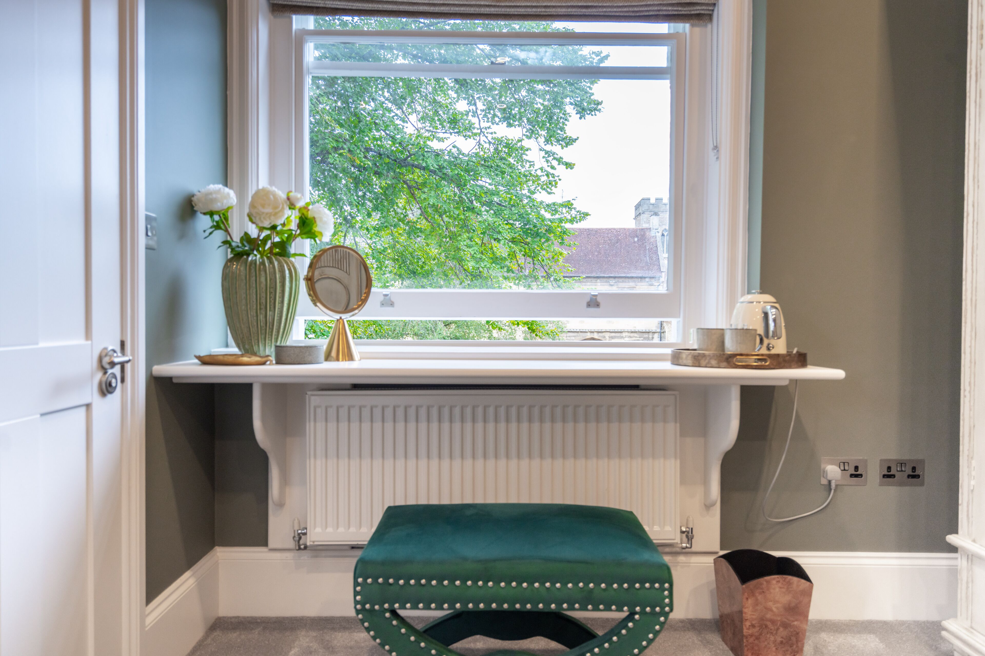Ett badrum med ett fönster, en radiator, en grön pall och en vas med blommor.
