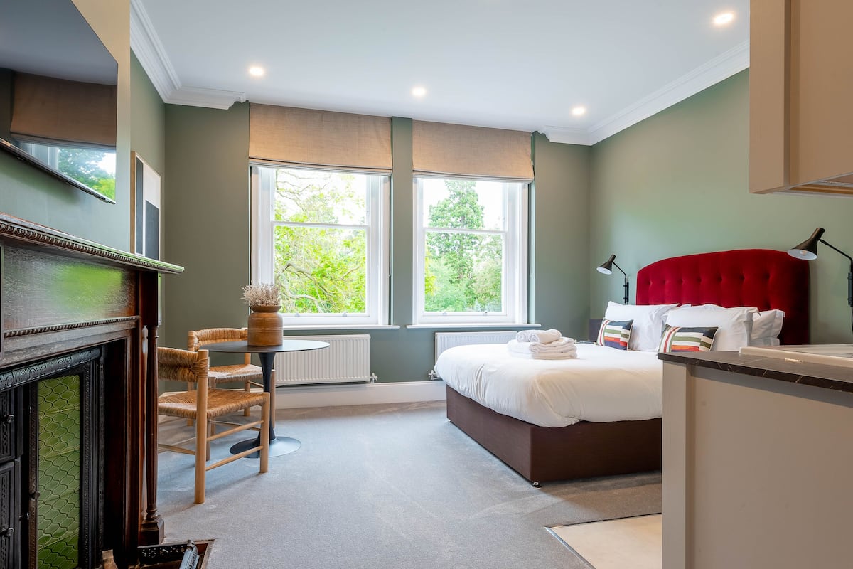 Een slaapkamer met een bed, een stoel, een klein tafeltje, een open haard en grote ramen.