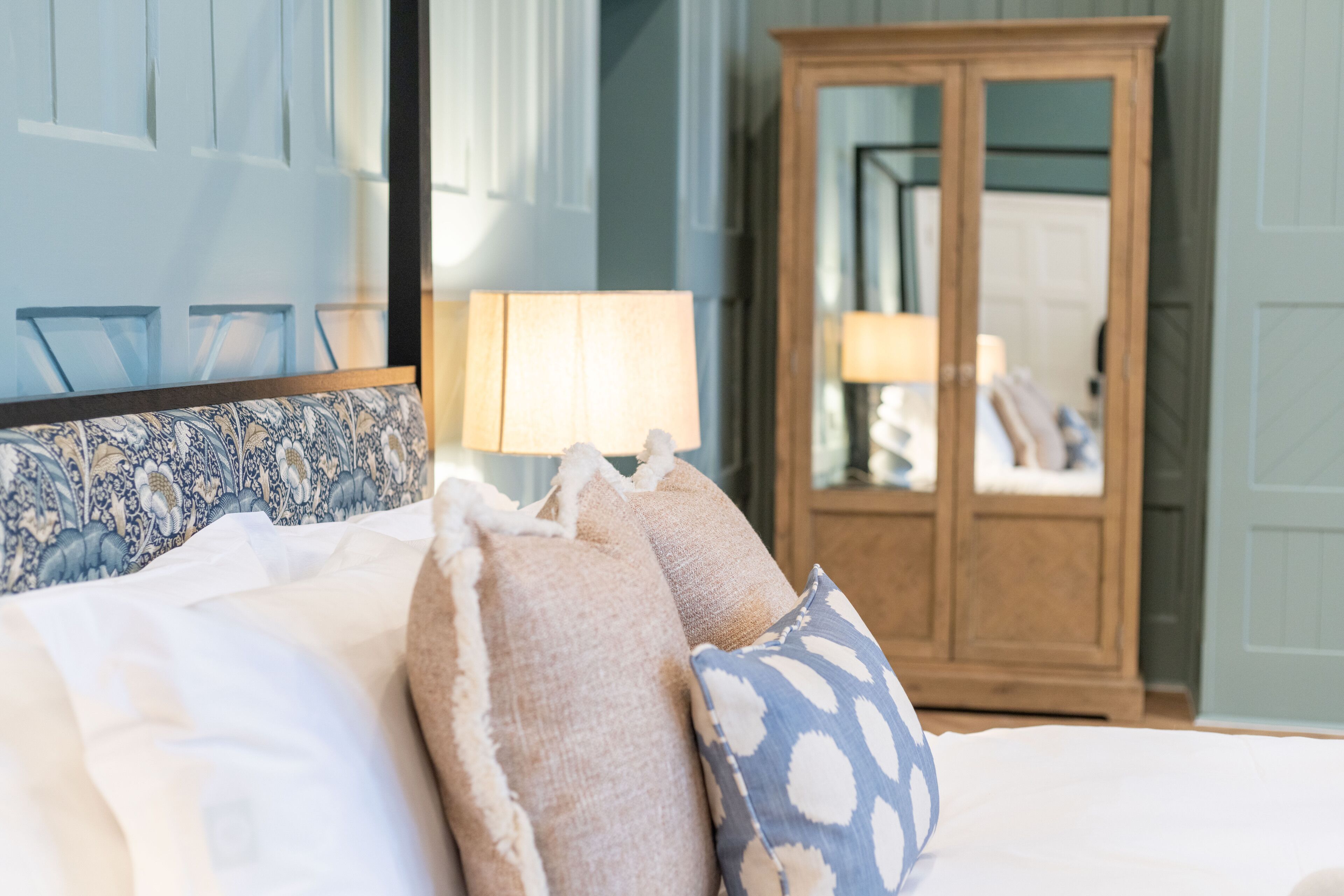 Una cama con ropa blanca y almohadas decorativas, un armario de madera y un espejo que refleja la habitación.