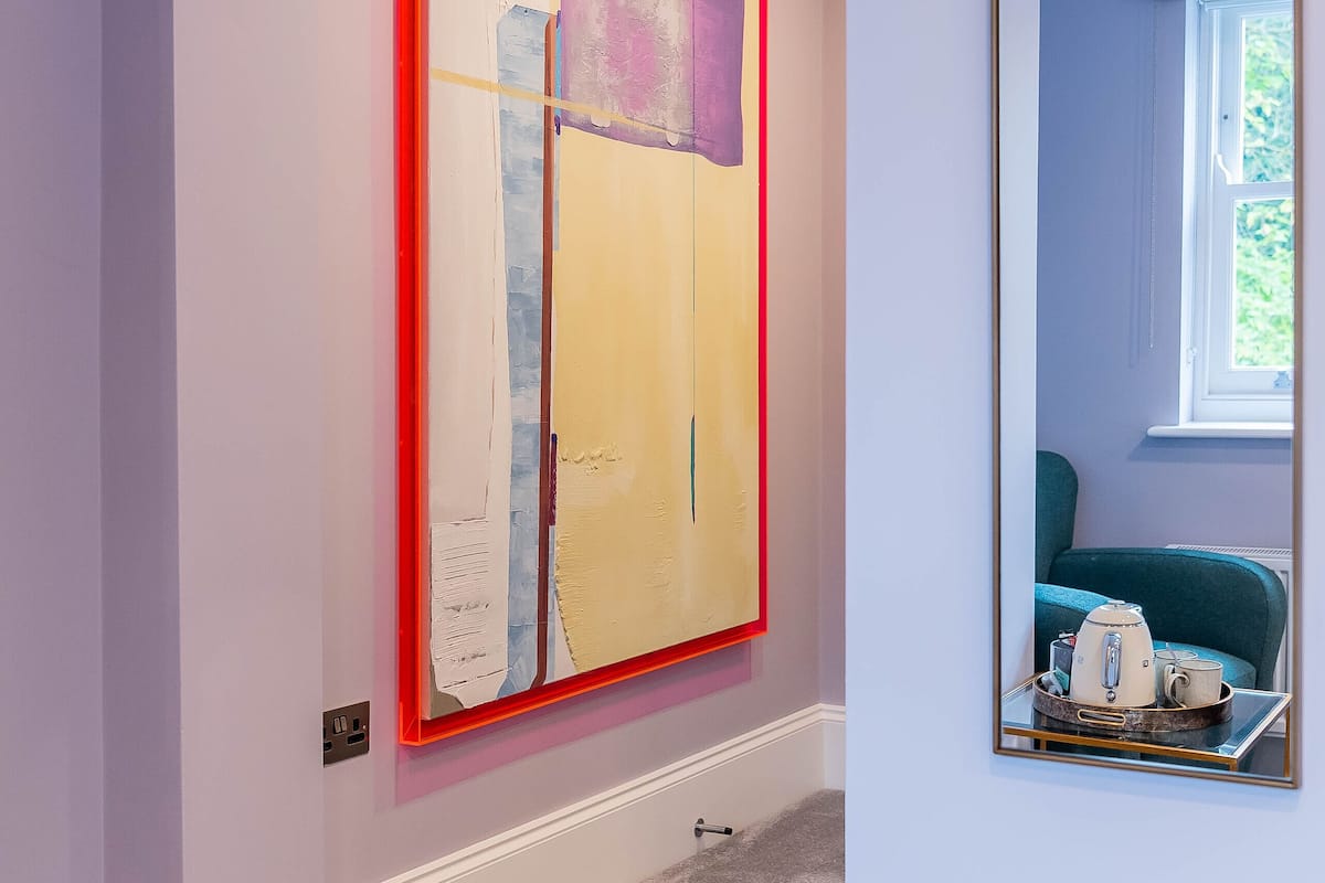 Een kamer met een groot abstract schilderij aan de muur, een spiegel en een raam met uitzicht op groen.