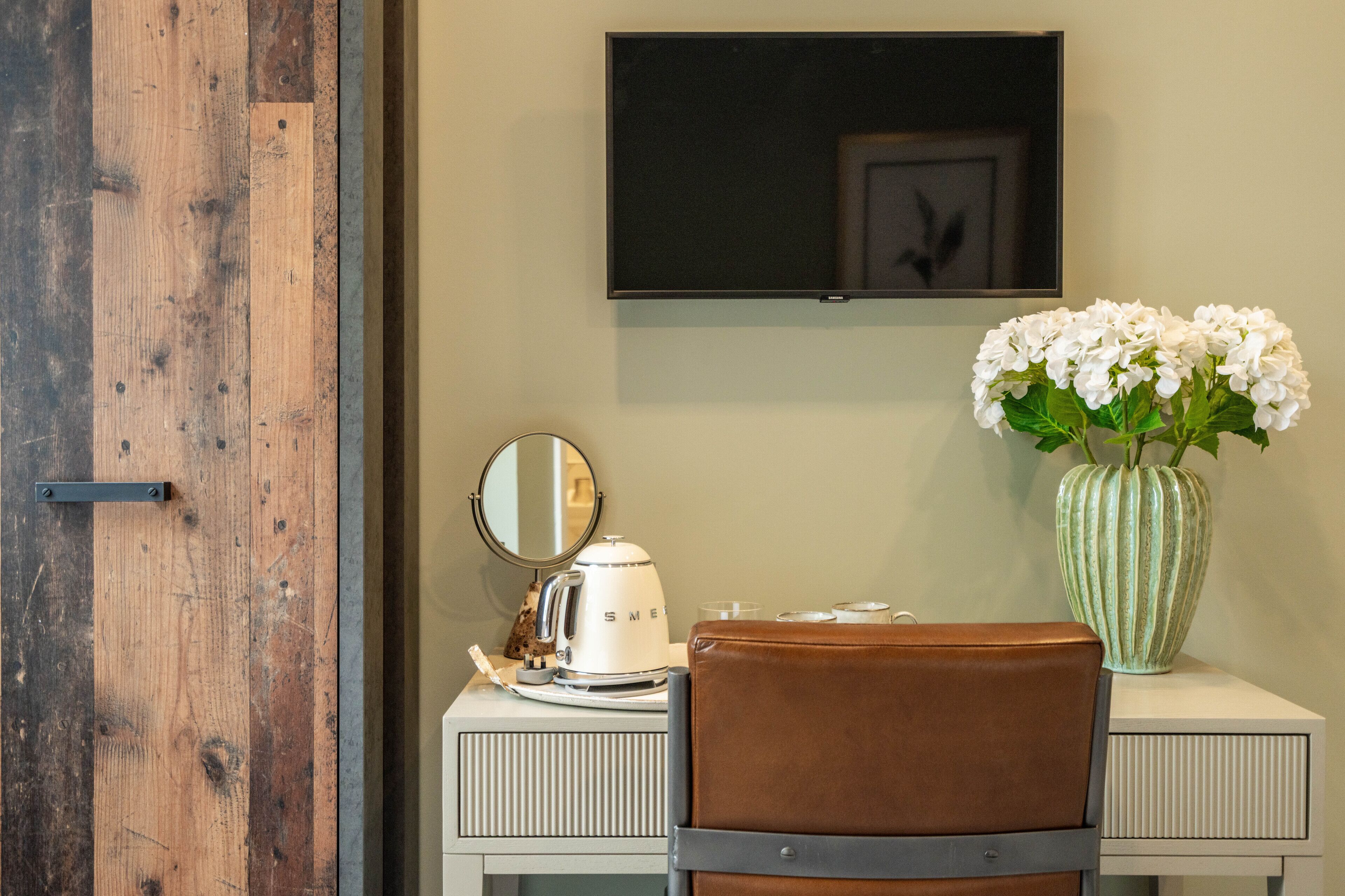A room with a wooden door, a white dresser, a brown leather chair, a white kettle, a mirror, a television mounted on the wall, and a vase with white flowers.