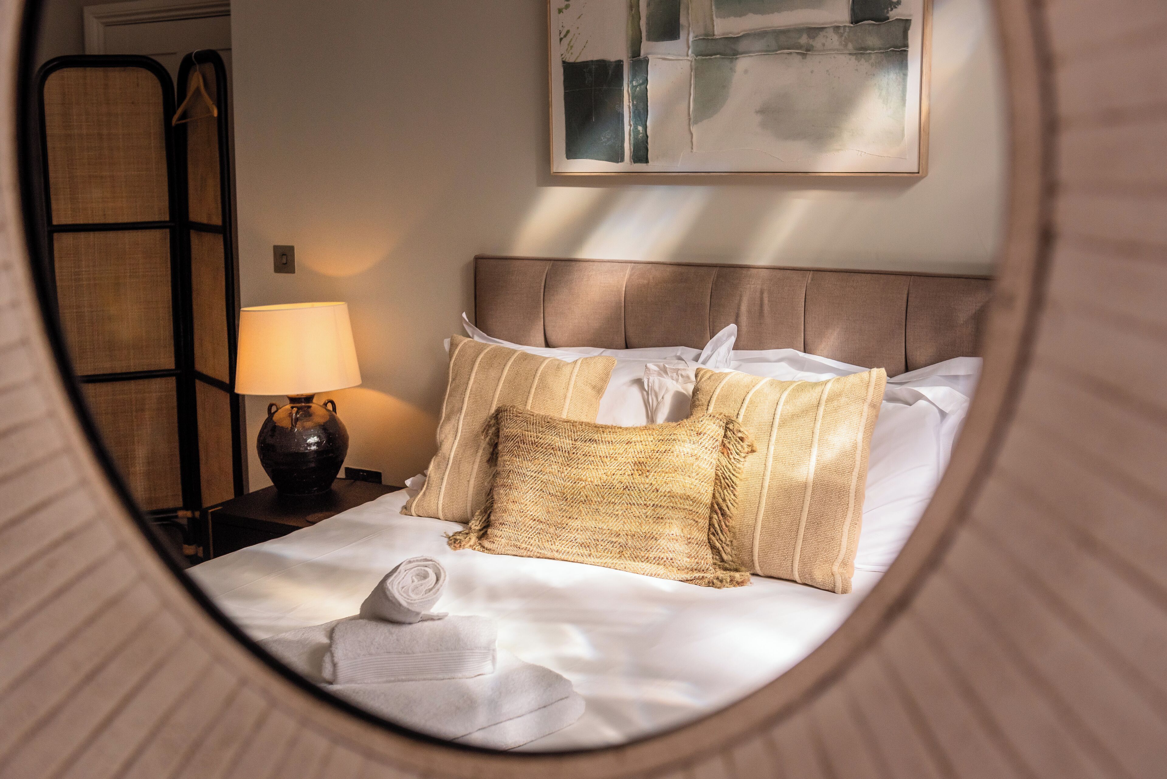 A neatly made bed with white linens and striped pillows, a bedside lamp, and a framed abstract painting on the wall.