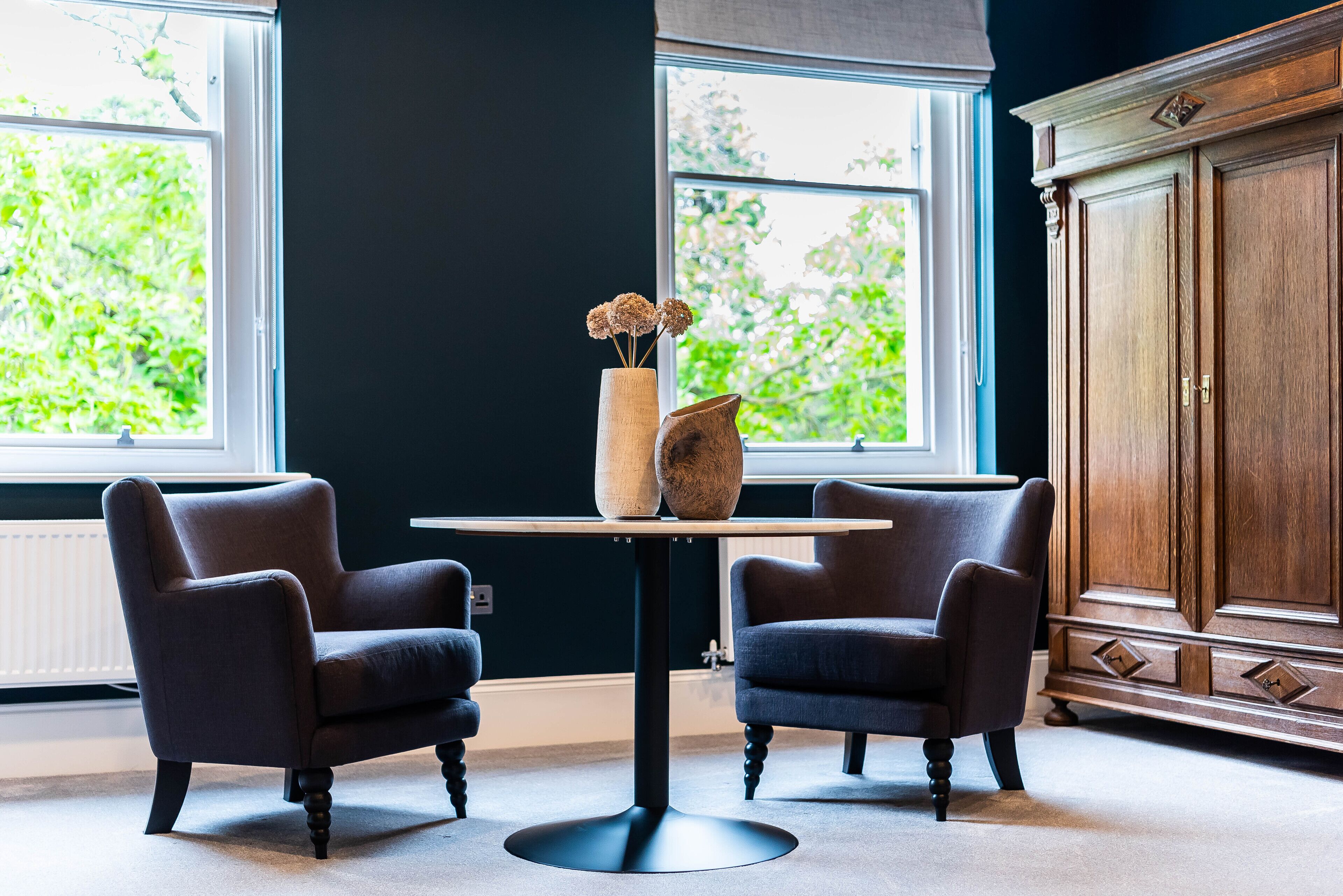 Um quarto com duas poltronas, uma mesa com um vaso e um armário de madeira.