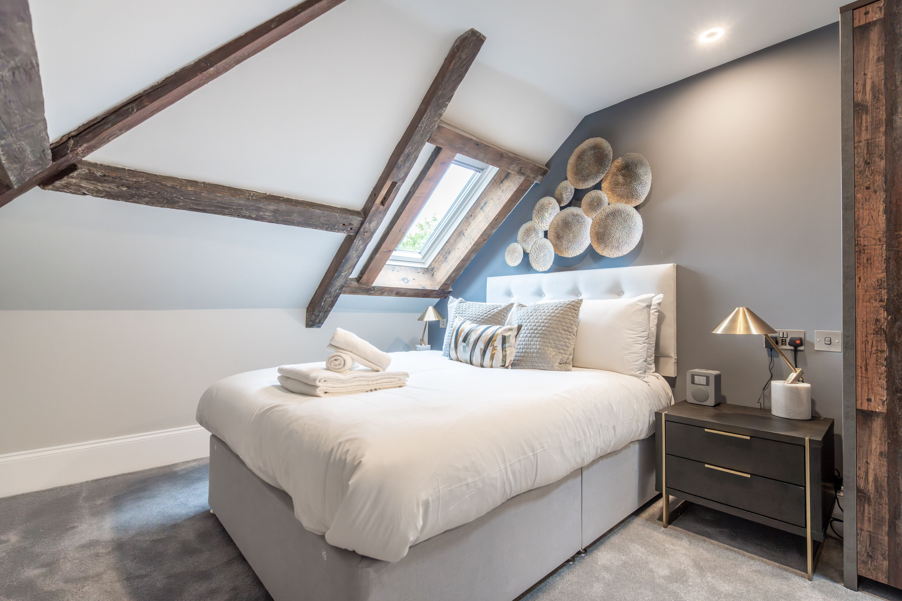 Un dormitorio con techo inclinado, una cama con ropa blanca, una mesita de noche con una lámpara y un objeto decorativo en la pared.