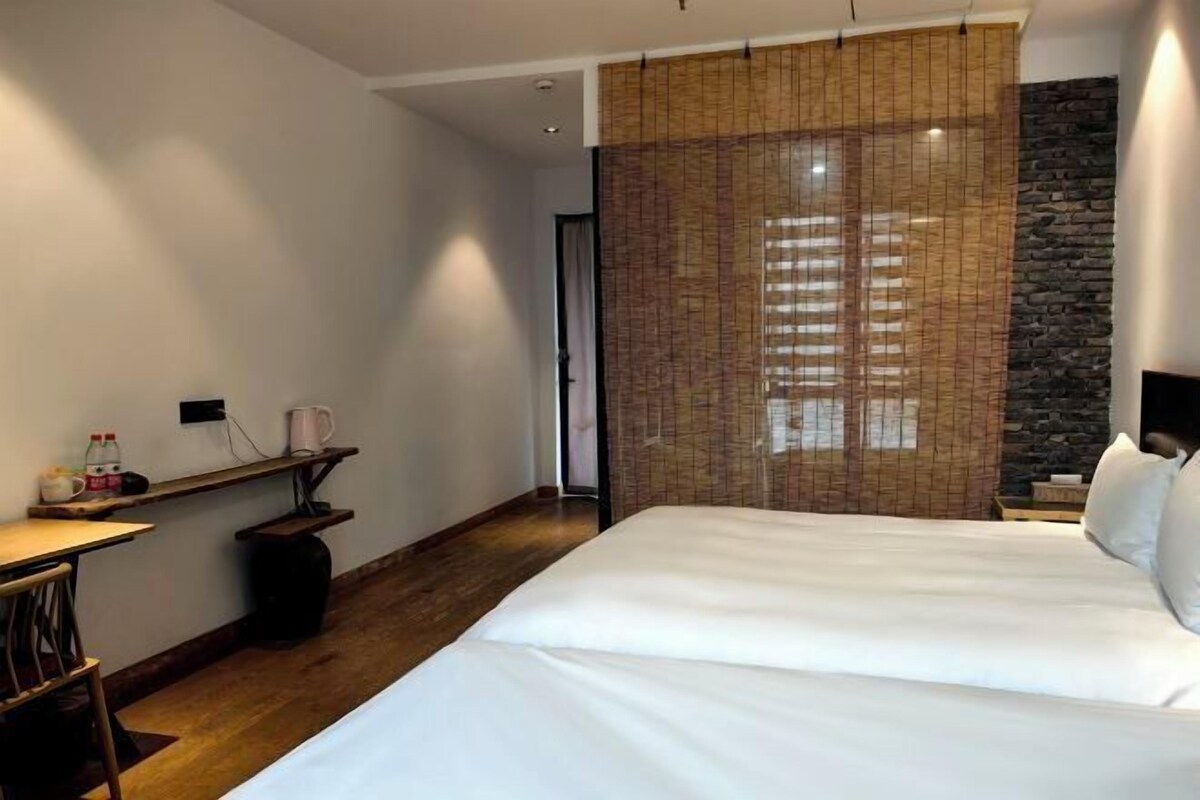 A hotel room with a large bed, a wooden desk, a small table, a stone accent wall, and a bamboo curtain.