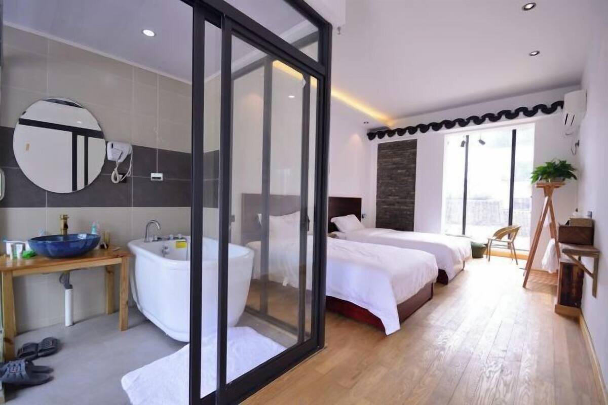 Un dormitorio moderno con una puerta de cristal que da al baño, un espejo redondo grande, una mesita de noche de madera, una bañera blanca y una cama con ropa de cama blanca.