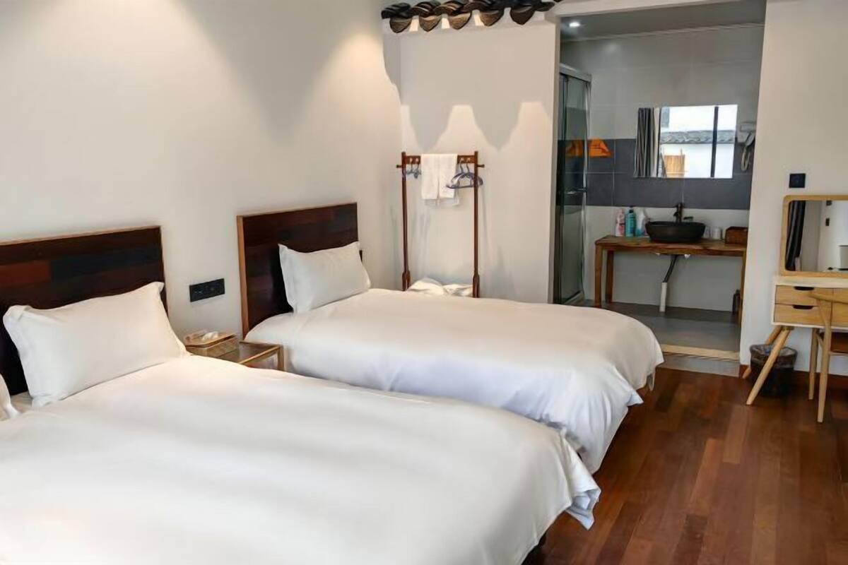 Habitación de hotel con dos camas, un escritorio y un baño visible a través de una puerta abierta.