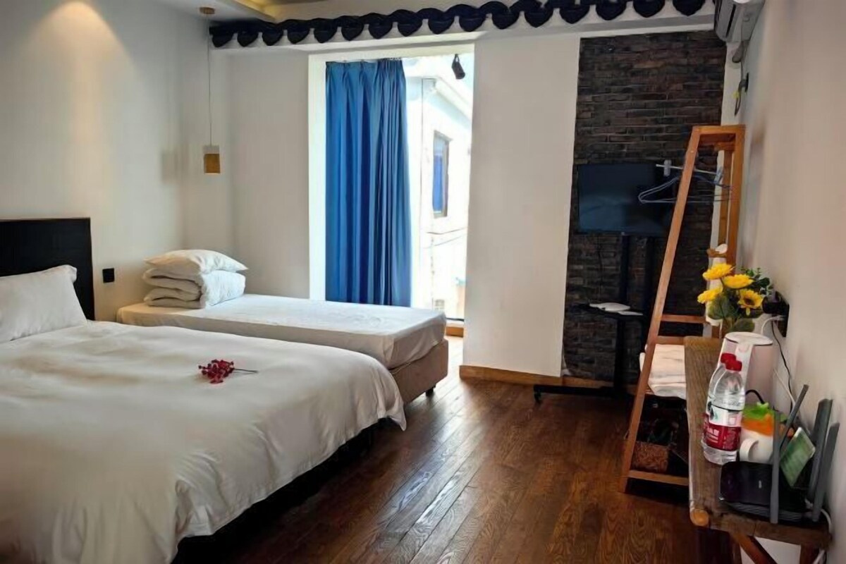 Quarto de hotel com duas camas, piso de madeira, parede de pedra, televisão e cadeira de madeira com uma garrafa e flores.