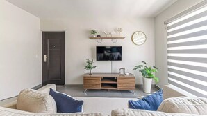 Una sala de estar moderna con una puerta oscura, un televisor de pantalla plana montado en la pared, un mueble de entretenimiento de madera y un ventanal con persianas.