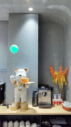 Une mascotte en forme d’ours tenant un plateau avec une pâtisserie, une machine à café et un panier de blé.
