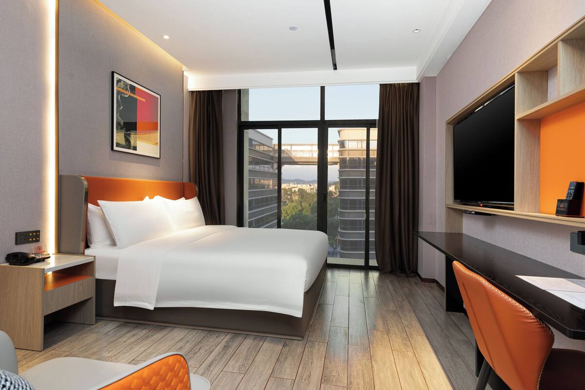 A modern hotel room with a large bed, a desk, a chair, a TV, and a window with a city view.