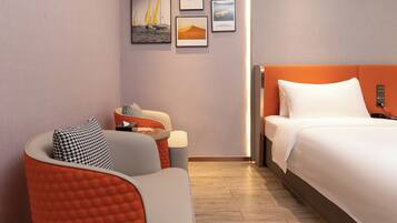 A modern hotel room with a bed, a seating area with a chair and ottoman, and three framed artworks on the wall.