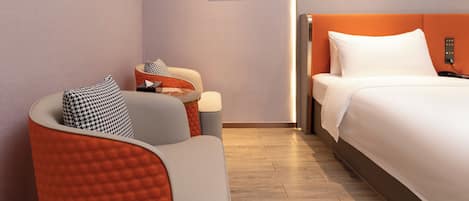 A modern hotel room with a bed, a seating area with a chair and ottoman, and three framed artworks on the wall.